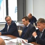 Grupa pieciu dorosłych, elegancko ubranych mężczyzn zasiadajacych przy biurkach na rozległej sali. Jeden z nich czyta z kartki i trzyma mikrofon.