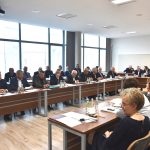 Grupa osób różnej płci i wieku zasiadający przy biurkach. Na ich blatach widać kartki papieru oraz wodę. Wszyscy znajdują się na rozległej sali.