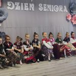 Grupa dzieci siedzi na ławce przebrana w długie kolorowe spódnice i chusty. Za nimi szara kurtyna z napisem Dzień Seniora.