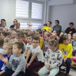 Grupa kilkudziesięciu osób, dzieci i starszych kobiet i mężczyzn siedzi na widowni w jasnej sali i ogląda przedstawienie dzieci.