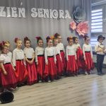 Grupa dziewczynek w czerownych dłuch spódnicach stoi w jasnej sali przedszkolnej. Za nimi szara kurtyna z napisem Dzień Seniora.