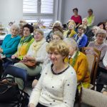 Grupa kilkudziesięciu osób starszych kobiet i mężczyzn siedzi na widowni w przedszkolu.