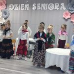 Grupa dzieci dziewczynek i chłopców przebranych w stroje długie spódnice i chusty stoi w sali przedszkolnej i recytuje wiersze. za nimi szara kurtyna z napisem Dzień Seniora.