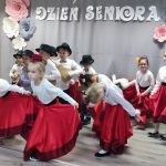 Grupa dziewczynek w długich spódnicach i białych bluzakach tańczy na sali przedszkolnej.