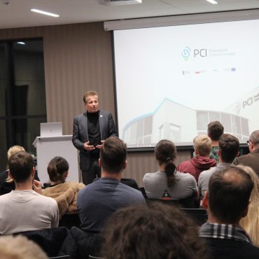 Starszy mężczyzna w garniturze stoi na sali pełnej ludzi i przemawia do publiczności. Za nim wyświetlją się slajdy na projektorze.