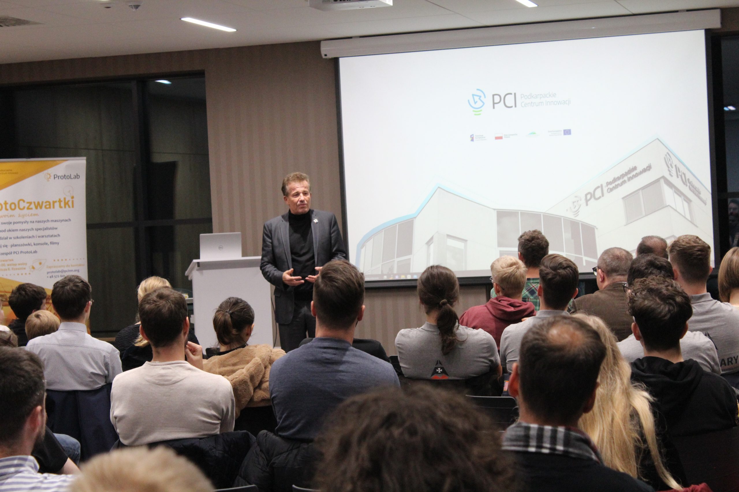 Starszy mężczyzna w garniturze stoi na sali pełnej ludzi i przemawia do publiczności. Za nim wyświetlją się slajdy na projektorze.