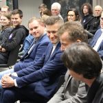 Grupa osób siedzi na sali i słucha mężczyzny przemawiającego.