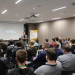 Grupa osób siedzi na sali i słucha starszego mężczyzny w garniturze. Za nimi ekran.