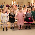 Znacząca grupa dzieci obojga płci stojaca na rozległej, dobrze oświetlonej sali, przy długich ławkach. Z tyłu widać grupę osób dorosłych.