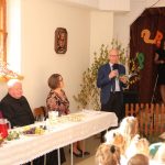 Grupa gości rożnej płci i wieku siedząca przy podłużnym stole na rozległej, dobrze oświetlonej sali. Jeden z gości (starszy mężczyzna w garniturze) przemawia za pomocą mikrofonu do zebranych.