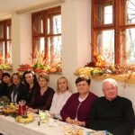 Grupa gości rożnej płci i wieku siedząca przy podłużnym stole na rozległej, dobrze oświetlonej sali.