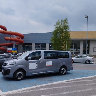 Bus koloru szarego stoi na miejscu parkingowym oznaczonym na niebiesko przed dyżym budynkiem.