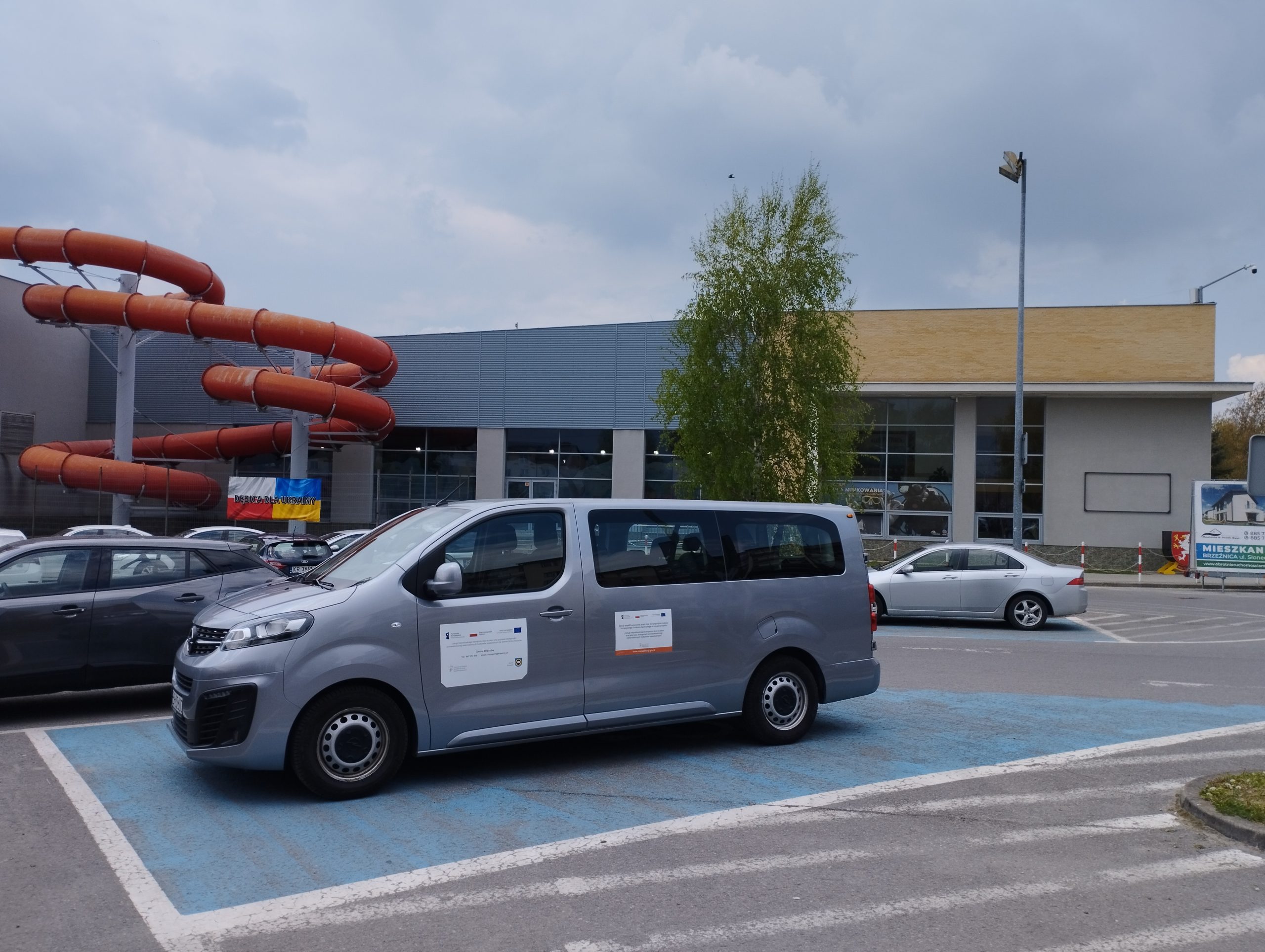 Bus koloru szarego stoi na miejscu parkingowym oznaczonym na niebiesko przed dyżym budynkiem.
