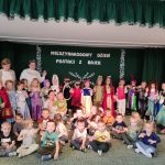 Grupa dzieci w strojach przedstawiających postacie z bajek oraz nauczycielki i kilkanaście dzieci w normalnych, codziennych ubraniach. Pozują do zdjęcia w grupie na jasnym dywanie. W tle widać ciemną kotarę.