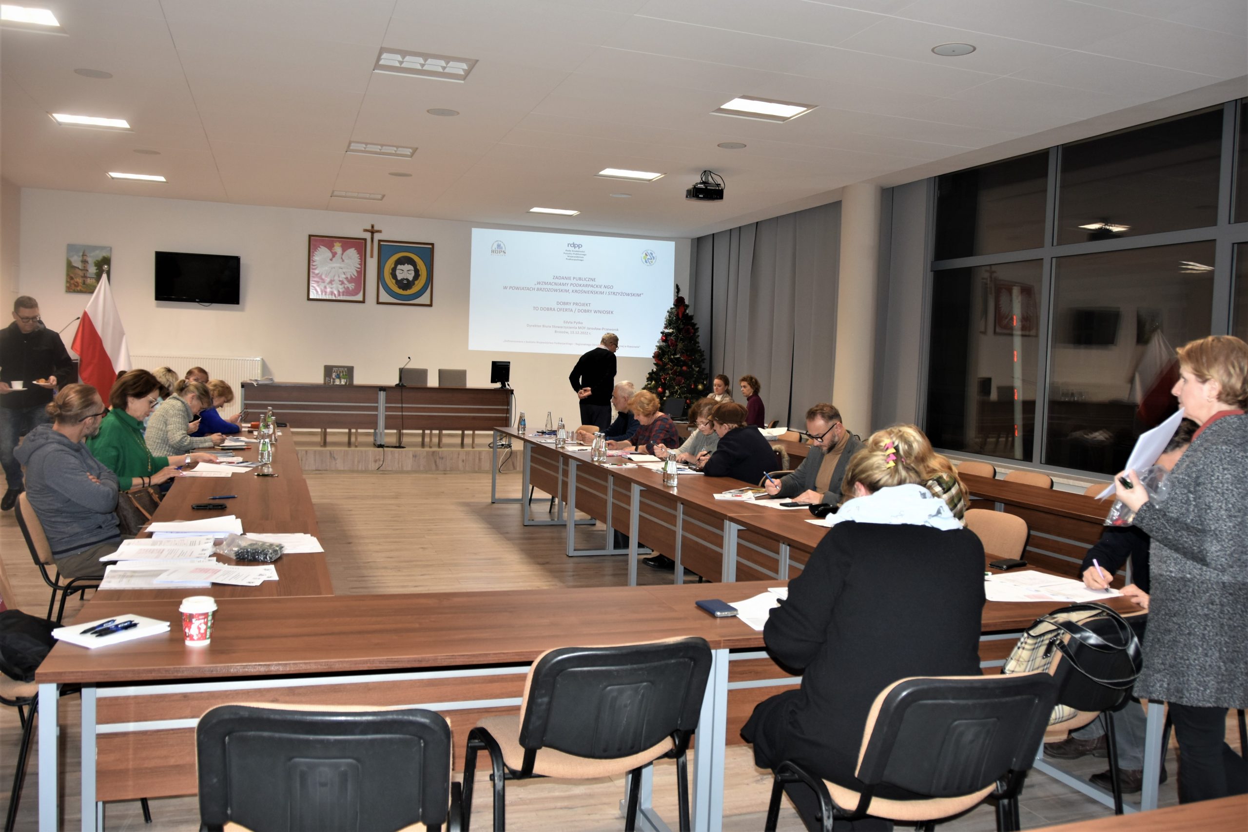 Grupa osób siedzi przy stolikach i słucha mężczyzny który prowadzi preleckję.