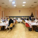 Grupa osób, gównie starszych siedzacych przy stołach na rozległej sali. Na stołach znadują się naczynia i sztućće.