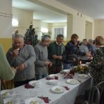 Grupa osób róznej płci i wieku dzieląca się opłatkiem przy stołach na duzej sali.