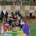 Grupa młodych szachistów na sali gimnastycznej w trakcie rozgrywek. W te widać grupę dorosłych i młodzieży.