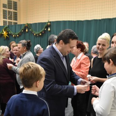 Grupa osób różnej płci i wieku dzieląca się opłatkiem na rozległej sali.