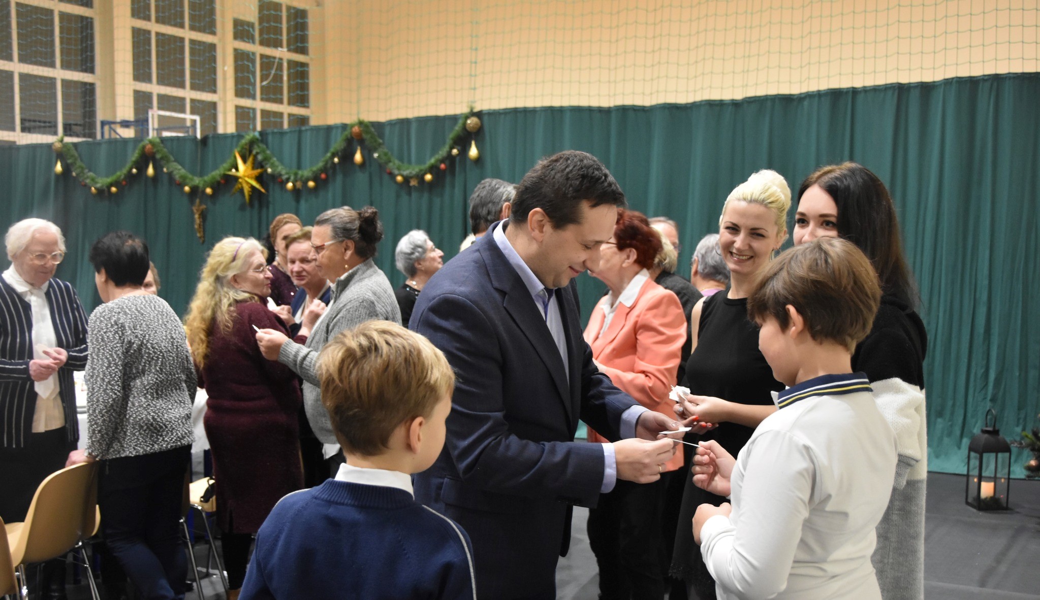 Grupa osób różnej płci i wieku dzieląca się opłatkiem na rozległej sali.