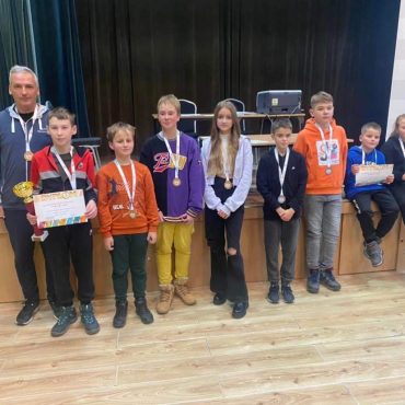 Grupa dzieci z dorosłym opiekunem pozująca do fotografi na rozległej sali. Dzieci posiadają medale, puchary i dyplom.