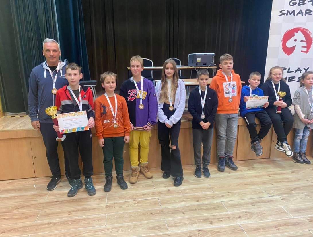 Grupa dzieci z dorosłym opiekunem pozująca do fotografi na rozległej sali. Dzieci posiadają medale, puchary i dyplom.