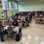 Grupa dzieci różnej płci i wieku grajaca w szachy na rozleglej sali. W tle widać trzy duże lustra.