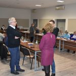 Grupa osób rozmawia w sali konferencyjnej. Za nimi siedzą ludzie przy długich stołach.