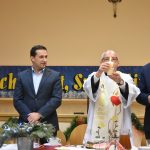 Trzech męzczyzn w graniturach oraz kapałan trzymajacy świece. Wszyscy stoją przed stołem.