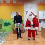 Mężczyzna w ciemnym płaszczu wraz z mężczyzną przebraną w czerwony strój mikołaja wchodzi do sali przedszkolnej. Mężczyzna trzyma w rękach w klocki.