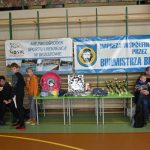 Grupa osób głównie dzieci na rozległej sali. Widac stoły z pucharami i nagrodami oraz dwa banery promocyjne.