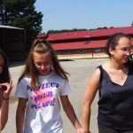 Trójka młodych dziewczyn w luźniejszych strojac oraz młody mężczyzna w jasnej koszuli. Z tyłu widać większy budynek.