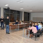 Grupa osób siedzi przy stolikach i słucha mężczyzny który prowadzi preleckję.