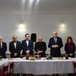 Grupa osób, głównie męzczyzn w graniturach i jedna kobieta stojacych przed długim stołem. Na stole są placki i potrawy.