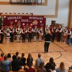 Grupa dzieci w strojach odświętnych i ludowych występująca na sali gimnastycznej. Obserwuje ich grupa dorosłych.