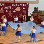 Grupa dziewczynek w białych koszulkach i błękitnych spódnicach występująca z dużą chusta na sali gimnastycznej.