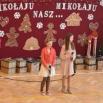 Dwie młode dziewczynki w odświętnych strojach występujące na scenie. W tle ścianka ze zdobieniami świątecznymi z napisem Mikołaju, Mikołaju Nasz.