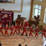 Grupa dzieci w stroju elfów i dorosły mężczyzna w stroju Mikołaja tańczący przed grupa osób na sali gimnastycznej.