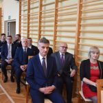 Grupa dorosłych mężczyzn i jedna kobieta w oficjalnych strojach siedzący na sali gimnastycznej.