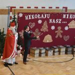 Mężczyzna w garniturze oraz mężczyzna w stroju Mikołaja na sali gimnastycznej. Obok nich lezą przygotowane prezenty.