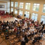 Grupa osób różnej płci i wieku zasiadający na krzesełkach w sali gimnastycznej. Wszyscy spoglądają na mikołajkowy występ.