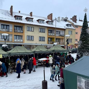 Grupa osób ogląda stoiska jarmarków na rynku. Panuje zima. W tle widać bloki.