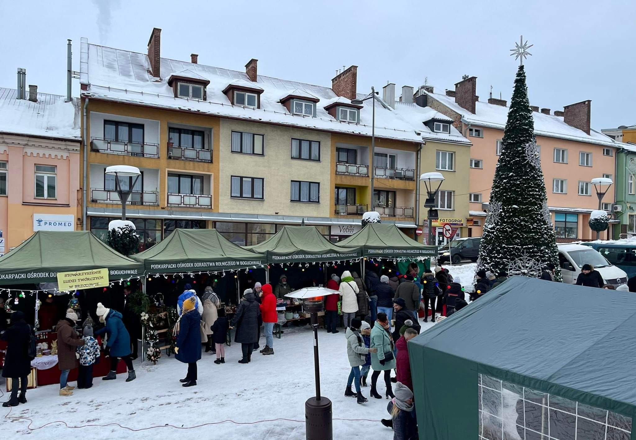 Grupa osób ogląda stoiska jarmarków na rynku. Panuje zima. W tle widać bloki.