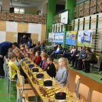 Grupa osób różnej płci i wieku na rozległej sali w trakcie rozgrywek szachowych. Na ścianie widać cztery banery.