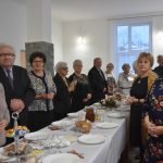 Grupa osób starszych stojąca przy podłużnym stole. Na stole znajduja się przystawki i zastawa.