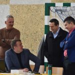 Rozmawiających czterech mężczyzn. Znajdują się na rozległej sali gimnastycznej. W tle widać bramkę.