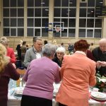 Grupa osób róznej płci i wieku dzieląca się opłatkiem przy stołach na duzej sali gminastycznej.