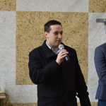 Dwoch dorosłych męzczyzn w oficjalnych storjach na sali gimnastycznej. Jeden z nich przemawia za pomocą mikrofonu.