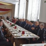 Grupa osób różnej płci siedzaca przy podłużnym stole na wielkiej sali. Na stole znajduje się zastawa oraz posiłki.