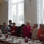 Grupa osób, głównie starszych dzieli się opłatkiem na duzej sali.